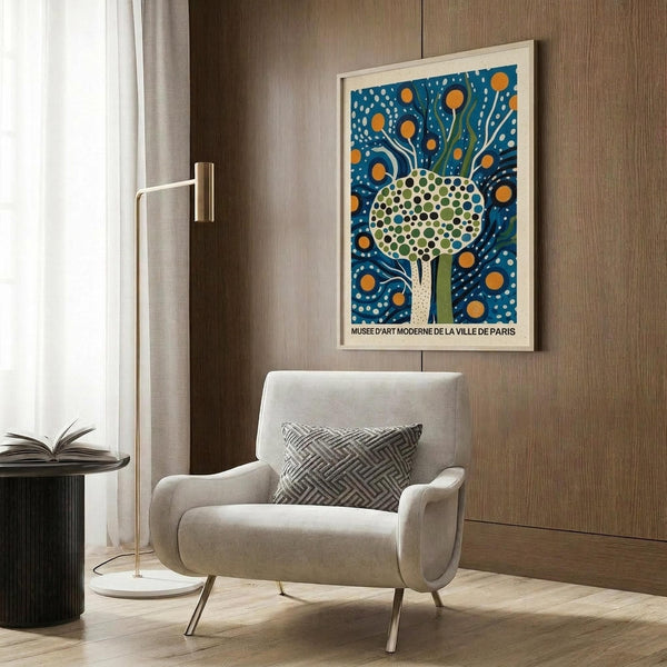 May include: Framed vintage-style art poster featuring stylized tree with dotted green canopy and flowing blue background with orange circles, displayed above beige upholstered armchair with geometric patterned pillow. Modern reading nook with brass floor lamp, dark round side table with open book, and warm wood paneled wall.