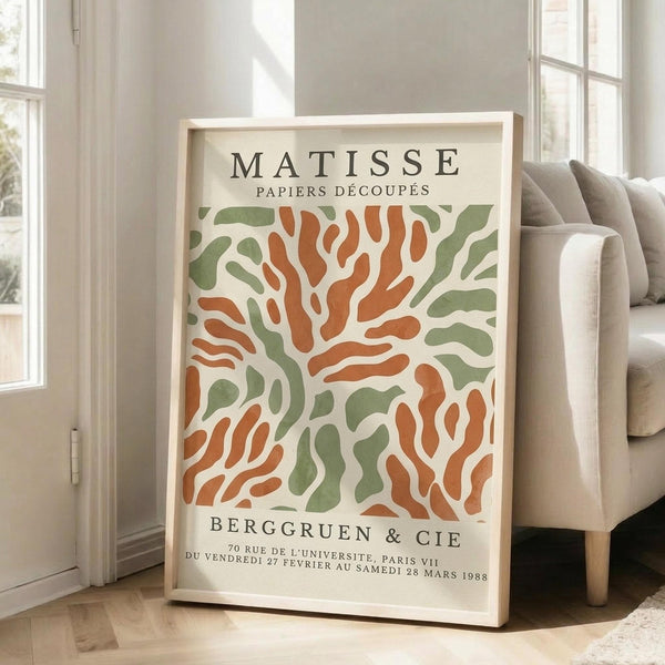 May include: Framed vintage-style exhibition poster featuring abstract organic shapes in terracotta orange and sage green on cream background, displayed on light wooden floors against white walls near a beige upholstered sofa. The artwork shows flowing, cut-paper inspired forms with French text at top and bottom, housed in a thin natural wood frame.