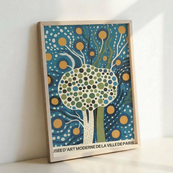 Framed art print featuring a stylized tree with a circular crown made of green, blue, and black polka dots against a deep blue background with swirling white lines and golden yellow circles. The tree has a white dotted trunk and the composition has a whimsical, modern folk art style. Text at bottom reads "MUSEE D'ART MODERNE DE LA VILLE DE PARIS" in black letters. The print is displayed in a light wood frame leaning against a white wall.