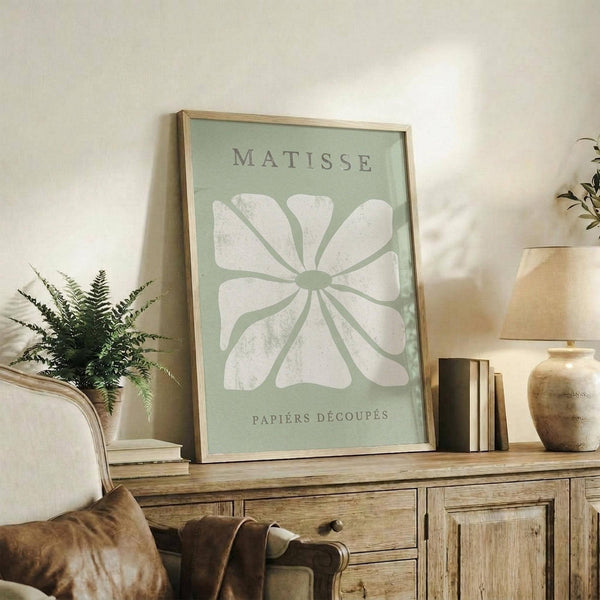 May include: Framed sage green poster with white abstract flower design and "Matisse Papiers Découpés" text, displayed on rustic wooden sideboard alongside fern plant, ceramic vase, and stacked books in warm-toned living room setting with cream upholstered chair