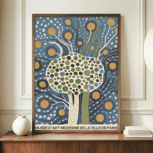 May include: Framed art print featuring a stylized tree with circular dotted foliage in green, blue and cream tones against a swirling blue background with golden orbs and white dots. The tree has flowing branches and trunk in white and green. Text at bottom reads "Musee d'Art Moderne de la Ville de Paris." Print displayed on wooden sideboard next to white ceramic vase and books in room with cream walls and white molding.