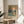 May include: Framed Monet-style winter landscape poster showing sailboat on water with snow-covered trees and orange sunset sky, displayed on round wooden side table next to beige upholstered chair, with plant leaves casting shadows on white wall in bright natural light