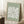 May include: Framed art print with sage green background featuring a white textured flower design in Matisse's paper cut-out style, displayed on white fluffy rug against white wall corner with natural sunlight, accompanied by wooden side table holding white coffee cup and saucer, with beige knit throw blanket visible at edge of frame.