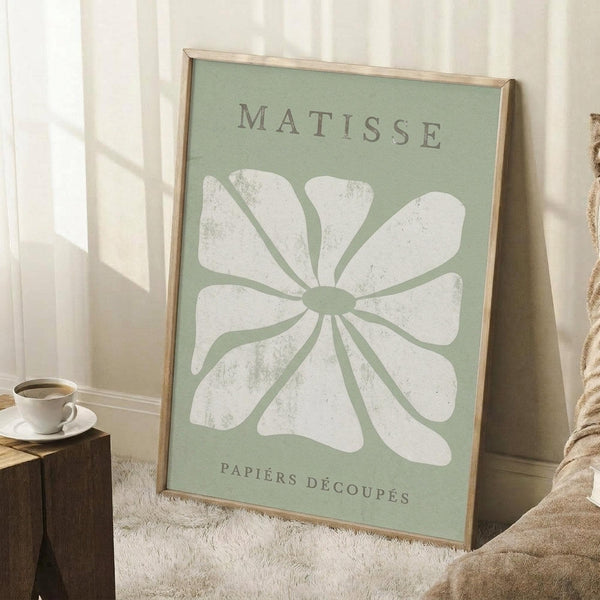 May include: Framed art print with sage green background featuring a white textured flower design in Matisse's paper cut-out style, displayed on white fluffy rug against white wall corner with natural sunlight, accompanied by wooden side table holding white coffee cup and saucer, with beige knit throw blanket visible at edge of frame.
