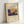 May include: Framed art print leaning against white wall showing abstract expressionist painting with vibrant blues, yellows, and rainbow arcs. Features geometric yellow mountain-like form, blue rectangular shapes, and flowing curved lines in warm colors. Natural wood frame on cream colored paper with cursive text above artwork and artist name below.