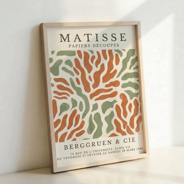 May include: Framed exhibition poster featuring Matisse's cut-out paper style artwork with flowing organic shapes in orange and sage green on cream background. Light wood frame displays the vintage 1988 Berggruen & Cie gallery poster, leaning against white wall with soft natural lighting casting gentle shadows.