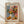 May include: Framed art poster in light wood frame leaning against white wall on wooden floor. Artwork shows colorful still life with geometric patterned vase containing flowers, oranges arranged nearby, painted in Matisse-inspired style with blue and pink background sections. Text reads "MATISSE" at top and "ART POSTER" at bottom. Natural lighting creates soft shadows on white fabric backdrop.