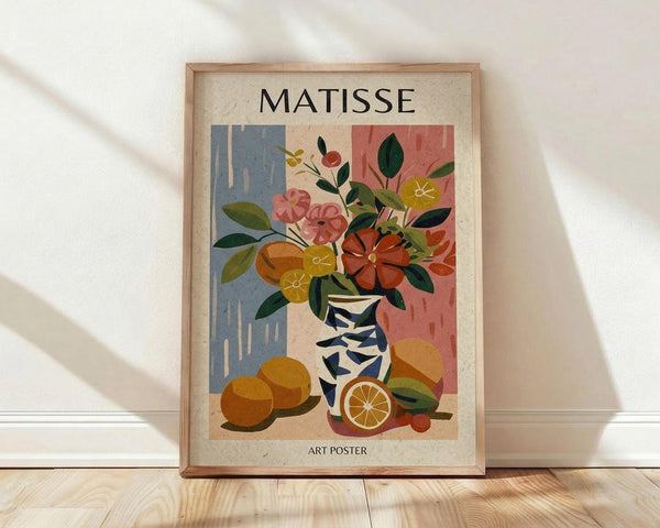 May include: Framed art poster in light wood frame leaning against white wall on wooden floor. Artwork shows colorful still life with geometric patterned vase containing flowers, oranges arranged nearby, painted in Matisse-inspired style with blue and pink background sections. Text reads "MATISSE" at top and "ART POSTER" at bottom. Natural lighting creates soft shadows on white fabric backdrop.