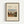 Framed vintage-style art print featuring Henri Rousseau's painting of Parisian outskirts with small figures walking through a village scene of houses, trees, and cloudy sky. The cream-colored poster displays black text reading "OUTSKIRTS OF PARIS" and "Henri Rousseau" with publication details. Set in a thin wooden frame with black matting against a white wall.