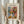 May include: Person holding framed art poster featuring colorful still life with flowers in geometric vase, oranges, and fruit arranged in Matisse-inspired style with blue and coral background blocks. Light wood frame against neutral curtained interior with dried pampas grass.