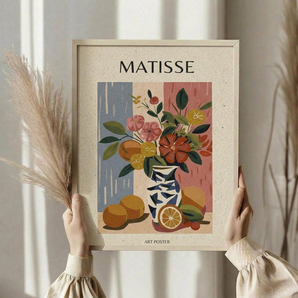 May include: Person holding framed art poster featuring colorful still life with flowers in geometric vase, oranges, and fruit arranged in Matisse-inspired style with blue and coral background blocks. Light wood frame against neutral curtained interior with dried pampas grass.