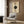 May include: Modern abstract geometric poster with cream background featuring three circles - two beige/brown half-circles at top and bottom, large black circle with radiating lines in center, and dotted horizontal line through middle. Text reads "MODERN" and "BAUHAUS" on sides. Shown framed on wood panel wall above beige upholstered armchair with patterned pillow, next to brass floor lamp and side table with open book.