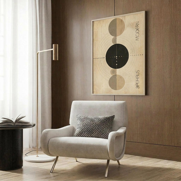 May include: Modern abstract geometric poster with cream background featuring three circles - two beige/brown half-circles at top and bottom, large black circle with radiating lines in center, and dotted horizontal line through middle. Text reads "MODERN" and "BAUHAUS" on sides. Shown framed on wood panel wall above beige upholstered armchair with patterned pillow, next to brass floor lamp and side table with open book.