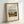 May include: Framed art print featuring Henri Rousseau's pastoral painting of a French village street scene with small figures walking along a dirt road between colorful houses under a cloudy blue sky. The cream-colored poster has black text reading "Outskirts of Paris" and artist information. Displayed in a thin light wood frame with black matting, leaning against a white wall.