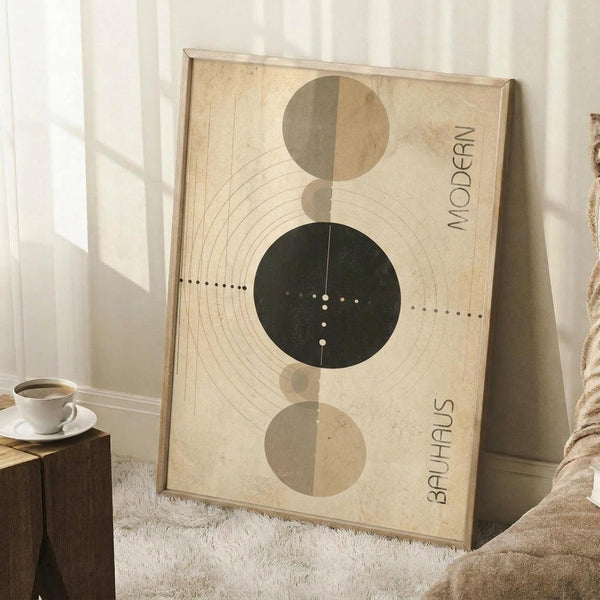 May include: Modern geometric art print in wooden frame leaning against white wall corner. Features abstract circles in sage green, beige, and cream with concentric line patterns and dotted details on textured paper background. Text reads "MODERN" and "BAUHAUS" vertically. Scene includes wooden side table with white coffee cup, beige throw blanket, and white textured rug in bright natural lighting.