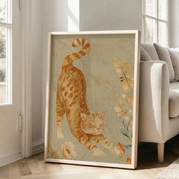 May include: Framed watercolor-style artwork featuring an orange tabby cat in a stretching pose against a soft beige background with delicate floral elements. The cat displays warm ginger and cream striped markings. Art is displayed in a light wooden frame leaning against a white wall next to a beige upholstered sofa in a bright, naturally lit living room with hardwood floors.