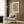 May include: Framed cat artwork displayed above a beige upholstered chair with geometric pillow in a modern living room setting. The print features an orange tabby cat stretching among delicate botanical elements in warm peachy tones with a light background and white mat in wooden frame. Scene includes brass floor lamp, side table with open book, and wood-paneled wall.