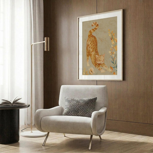 May include: Framed cat artwork displayed above a beige upholstered chair with geometric pillow in a modern living room setting. The print features an orange tabby cat stretching among delicate botanical elements in warm peachy tones with a light background and white mat in wooden frame. Scene includes brass floor lamp, side table with open book, and wood-paneled wall.