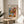May include: Framed poster with colorful still life artwork featuring flowers in a geometric vase alongside oranges, displayed on a round wooden side table next to a beige upholstered chair. Natural sunlight creates soft shadows on white walls. The artwork shows vibrant blues, pinks, oranges and greens in a modern artistic style with botanical elements.