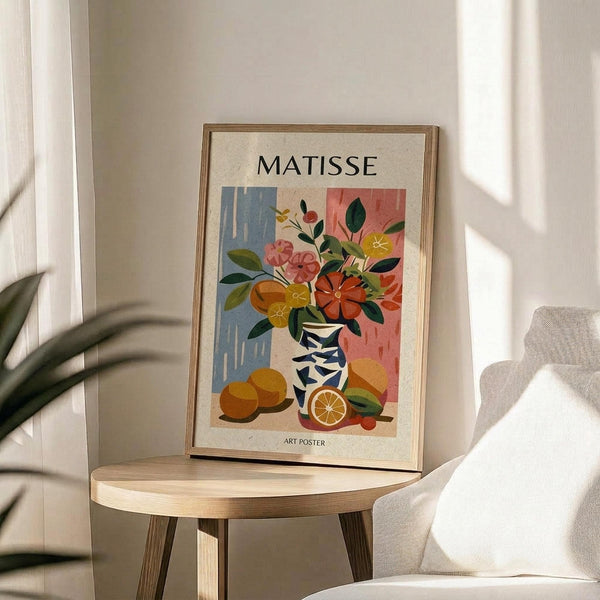May include: Framed poster with colorful still life artwork featuring flowers in a geometric vase alongside oranges, displayed on a round wooden side table next to a beige upholstered chair. Natural sunlight creates soft shadows on white walls. The artwork shows vibrant blues, pinks, oranges and greens in a modern artistic style with botanical elements.