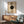 May include: Mid-century modern living room with sage green upholstered chair, wood sideboard, and ceramic table lamp. Framed geometric poster on white wall features beige and black circles with radiating lines and "MODERN" and "BAUHAUS" text. Warm natural lighting creates shadows across the minimalist Scandinavian-style interior with light wood furniture and neutral color palette.