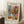 May include: A framed art poster leaning against a white wall featuring a colorful still life with flowers in a striped vase, oranges, and foliage in Matisse's cut-out style. The artwork displays vibrant blues, corals, greens, and oranges against a cream background. Scene includes a wooden side table with white teacup, textured cream rug, and soft natural lighting creating window shadows on the wall.