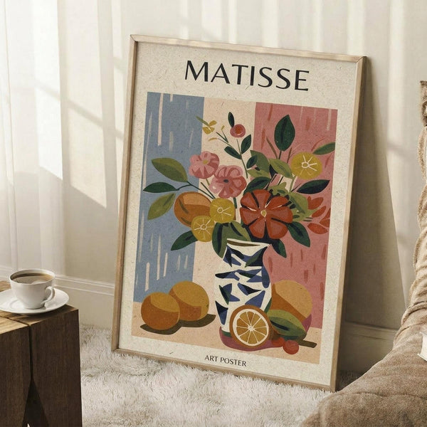 May include: A framed art poster leaning against a white wall featuring a colorful still life with flowers in a striped vase, oranges, and foliage in Matisse's cut-out style. The artwork displays vibrant blues, corals, greens, and oranges against a cream background. Scene includes a wooden side table with white teacup, textured cream rug, and soft natural lighting creating window shadows on the wall.