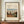 May include: Framed art print featuring Henri Rousseau's painting of a Parisian suburban street scene with houses, trees, and small figures under a cloudy sky. The cream-colored poster has vintage-style typography reading "Outskirts of Paris" and artist attribution. Displayed on wooden sideboard with white ceramic vase and books against cream paneled wall in modern interior setting.