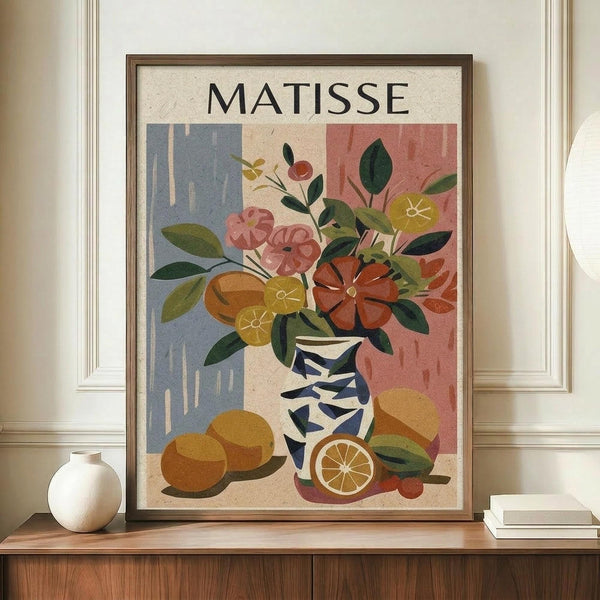 May include: Framed art print in wooden frame displaying colorful still life with flowers in blue and white patterned vase, oranges, and citrus slices against geometric blue and coral background. Print sits on wooden sideboard next to white ceramic vase and books in neutral-toned room with white paneled walls.