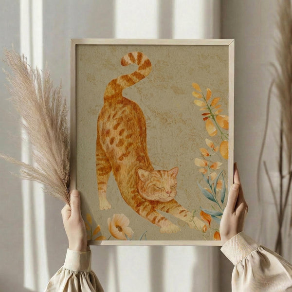 May include: Hands holding a framed watercolor artwork of an orange tabby cat stretching with its tail curled up, surrounded by delicate autumn-colored botanical elements including orange flowers and green stems. The painting has a soft beige background with a natural wood frame. Person wearing cream ruffled sleeves holds dried pampas grass in one hand while displaying the artwork against white curtains in natural lighting.