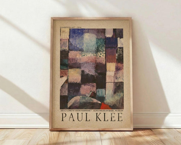 May include: Framed abstract artwork poster leaning against white wall on light wood flooring. The piece features geometric rectangular patches in muted colors including blues, purples, greens, pinks, and earth tones arranged in a mosaic-like pattern. Text reads "Exhibition Art - 1914" at top and "About a Motif from Hammamet - Kunstmuseum Basel Museum" and "Paul Klee" at bottom on beige background. Natural lighting creates soft shadows on the wall.