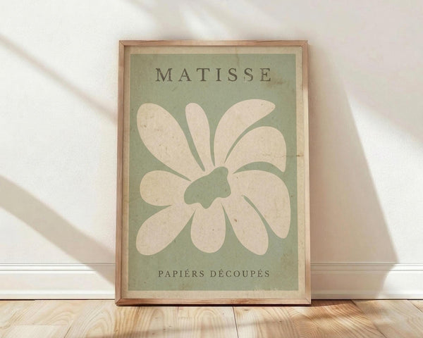 May include: Framed art print with sage green background featuring cream-colored abstract flower or leaf cutout design in Matisse's paper cutout style. Text reads "MATISSE" at top and "PAPIERS DÉCOUPÉS" at bottom. Natural wood frame sits on light wooden floor against white wall with soft window light creating shadows across the scene.