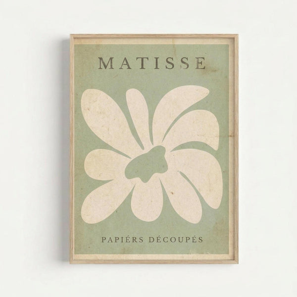 A framed poster featuring a large white flower or star shape with rounded petals on a sage green background. The word "MATISSE" appears at the top and "PAPIERS DÉCOUPÉS" at the bottom in dark text. The artwork is displayed in a simple light wood frame against a white wall, showing the cut-paper collage style characteristic of Matisse's later work with soft, organic forms.