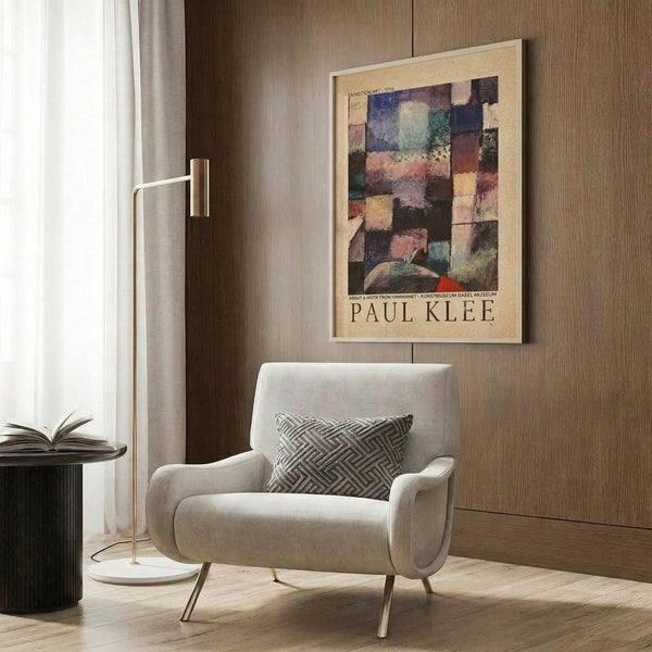 May include: Framed Paul Klee exhibition poster with abstract colorful geometric squares in blues, greens, oranges, and purples hanging on wood-paneled wall above beige upholstered chair with geometric patterned pillow, brass floor lamp, and dark round side table with open book in modern neutral living room setting