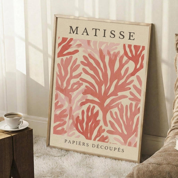 May include: Framed art print leaning against white wall corner next to coffee cup on wooden block and textured throw blanket. The poster features coral pink organic leaf-like cutout shapes on cream background with "MATISSE" at top and "PAPIERS DÉCOUPÉS" at bottom in black text. Natural wood frame, displayed on white fluffy rug with soft window lighting creating shadows on wall.