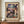 May include: Framed exhibition poster featuring Paul Klee's abstract artwork with colorful geometric squares in blues, purples, pinks, and earth tones. Poster displays on wooden sideboard next to white ceramic vase and stacked books against cream walls with white trim molding.