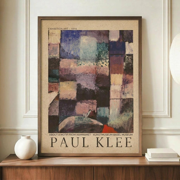 May include: Framed exhibition poster featuring Paul Klee's abstract artwork with colorful geometric squares in blues, purples, pinks, and earth tones. Poster displays on wooden sideboard next to white ceramic vase and stacked books against cream walls with white trim molding.