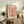 May include: Framed poster with coral-pink organic cutout shapes resembling seaweed or coral branches on cream background, displayed on rustic wooden sideboard alongside fern plant, ceramic vase, and books in warm neutral living room setting with beige armchair