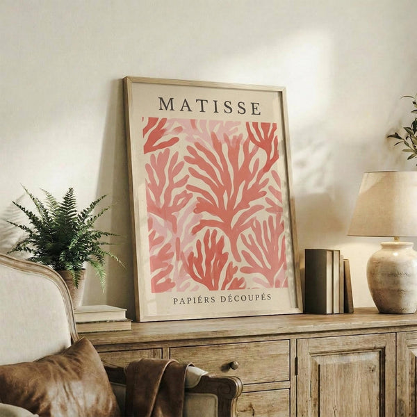 May include: Framed poster with coral-pink organic cutout shapes resembling seaweed or coral branches on cream background, displayed on rustic wooden sideboard alongside fern plant, ceramic vase, and books in warm neutral living room setting with beige armchair