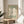 May include: Framed poster with sage green background featuring large white flower cutout design in Matisse's paper collage style, displayed on round wooden side table next to beige upholstered chair and plant leaves, with natural sunlight casting shadows on neutral wall