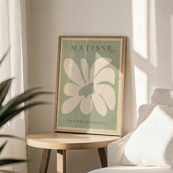 May include: Framed poster with sage green background featuring large white flower cutout design in Matisse's paper collage style, displayed on round wooden side table next to beige upholstered chair and plant leaves, with natural sunlight casting shadows on neutral wall