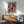 May include: Paul Klee abstract art poster with colorful geometric patches in blues, greens, pinks and earth tones, displayed in thin wooden frame above mid-century modern wood credenza. Sage green upholstered armchair with wooden arms sits nearby, white table lamp on credenza, natural sunlight streaming across white wall creating geometric shadows.