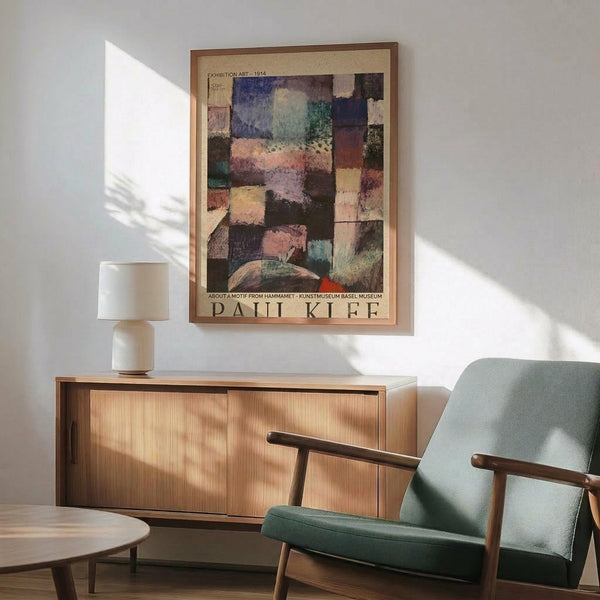 May include: Paul Klee abstract art poster with colorful geometric patches in blues, greens, pinks and earth tones, displayed in thin wooden frame above mid-century modern wood credenza. Sage green upholstered armchair with wooden arms sits nearby, white table lamp on credenza, natural sunlight streaming across white wall creating geometric shadows.