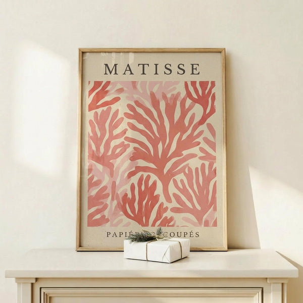 May include: Framed art print featuring coral pink cut-out leaf and botanical shapes in Matisse's signature paper collage style, displayed on light wood furniture next to a small wrapped gift with greenery. The poster has "MATISSE" at top and "PAPIERS DÉCOUPÉS" at bottom in black text, set in a thin wooden frame against cream colored wall.