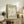 May include: Framed sage green poster with white flower cutout design in Matisse style displayed on rustic wooden sideboard. Scene shows neutral living room with fern plant, stacked books, ceramic vase, and beige upholstered chair. Warm natural lighting creates cozy modern farmhouse aesthetic with earth tone color palette.