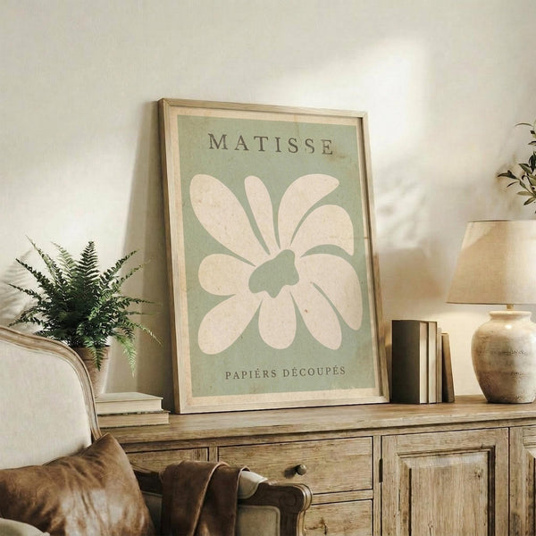 May include: Framed sage green poster with white flower cutout design in Matisse style displayed on rustic wooden sideboard. Scene shows neutral living room with fern plant, stacked books, ceramic vase, and beige upholstered chair. Warm natural lighting creates cozy modern farmhouse aesthetic with earth tone color palette.