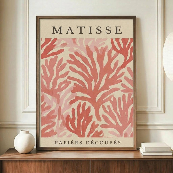 May include: Framed art print featuring coral-pink organic leaf-like shapes in Matisse's paper cut-out style against cream background, displayed on wooden sideboard next to white ceramic vase and stacked books in elegant room with white paneled walls