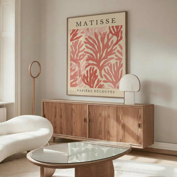 May include: Framed coral pink Matisse-inspired cut-out botanical poster displayed above a wooden sideboard in a modern living room. The artwork features organic leaf-like shapes in warm coral tones on cream background. Room shows white curved chair, glass oval coffee table, brass floor lamp, and small table lamp. Neutral walls and natural lighting create a mid-century modern aesthetic.