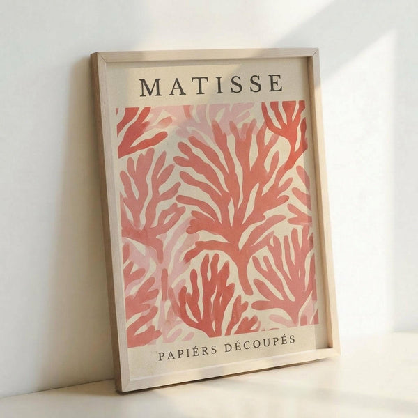 May include: Framed art print featuring coral-colored organic flowing shapes in Matisse's cut-out paper style against cream background, displayed in light wood frame leaning against white wall corner. Abstract botanical forms create dynamic movement across the composition with "MATISSE" and "PAPIERS DÉCOUPÉS" text at top and bottom.