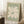 May include: Framed vintage-style art print with sage green background featuring large cream-colored abstract flower or leaf cutout design, displayed leaning against white wall corner on textured rug, with coffee cup on wooden block and beige throw blanket visible in cozy room setting with natural window light.